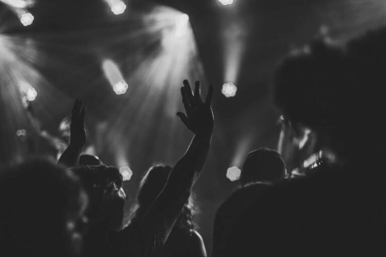 Silhouettes of people with hands raised during a worship service, with bright stage lights in the background.
