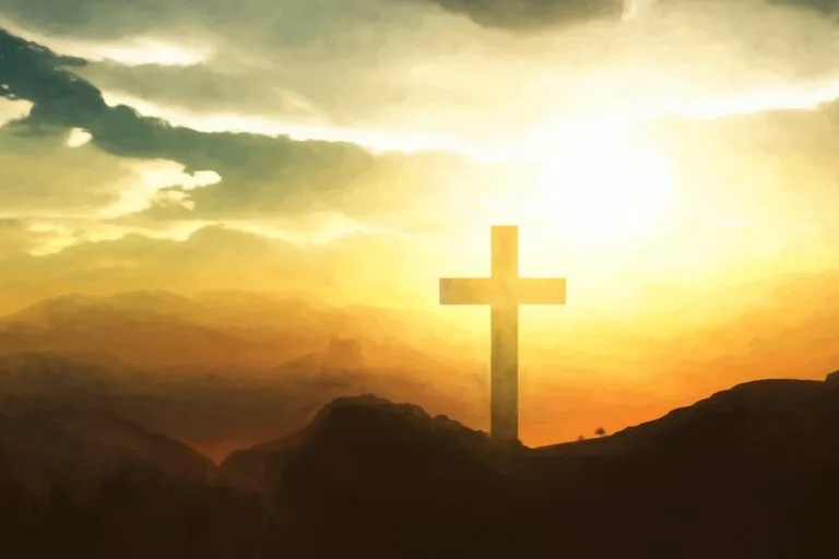 Cross on a hill silhouetted against a bright sunrise over distant mountains.
