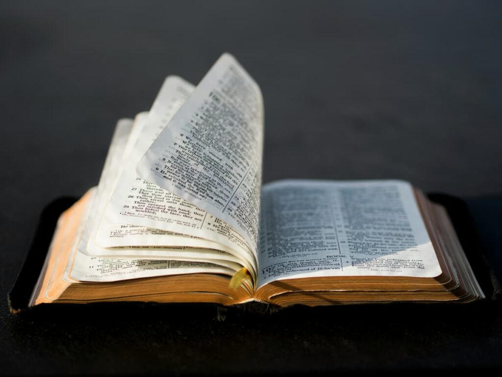 An open Bible rests on a dark surface, its thin pages curling upward and glowing softly in warm, low light.