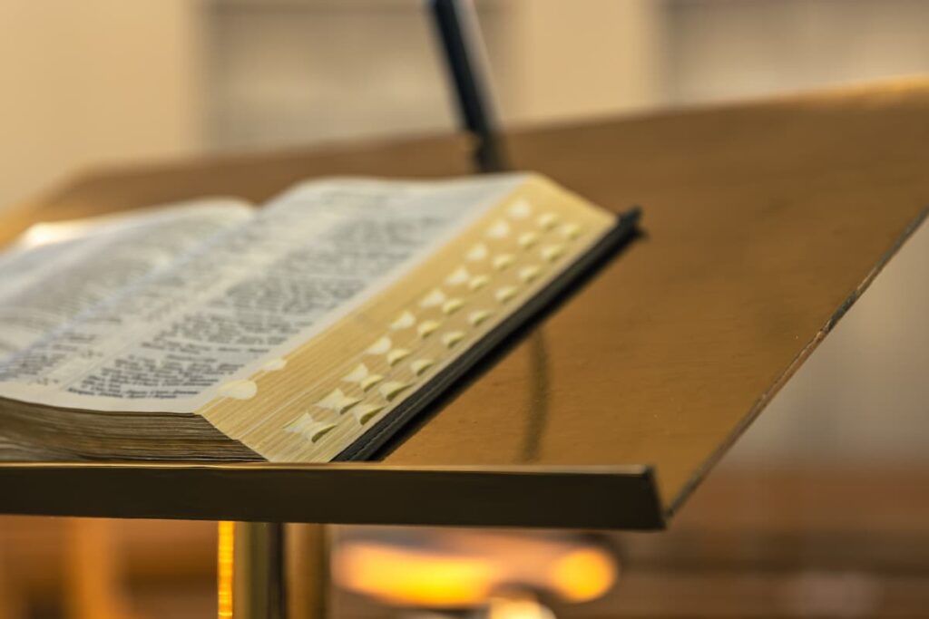 Open Bible with gold-edged thumb-index tabs resting on an angled wooden lectern, lit by warm bokeh light in a softly blurred sanctuary.