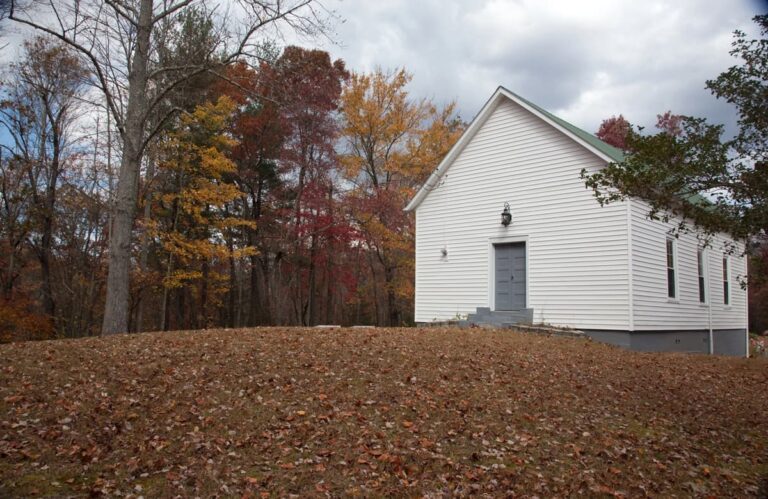 White clapboard one-room country church on a leaf-covered hillside, framed by trees in autumn color under an overcast sky.