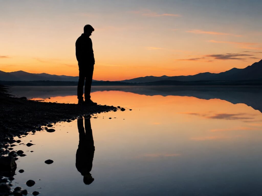 Silhouette of a man standing beside a lake looking at his reflection in the water.