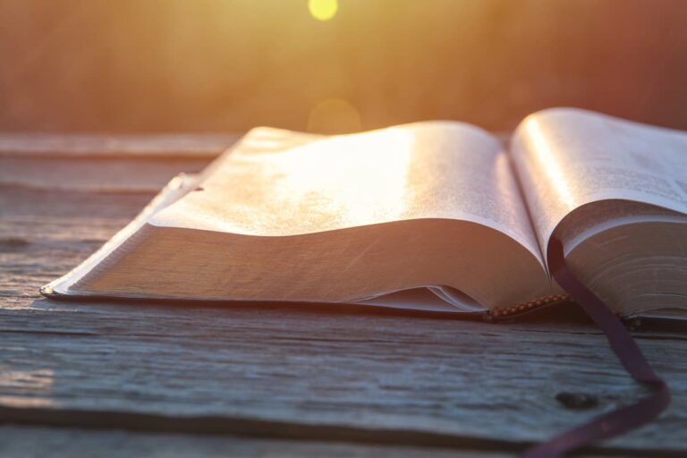An open Bible with a ribbon bookmark rests on a weathered wooden table, bathed in warm golden sunlight at sunset.